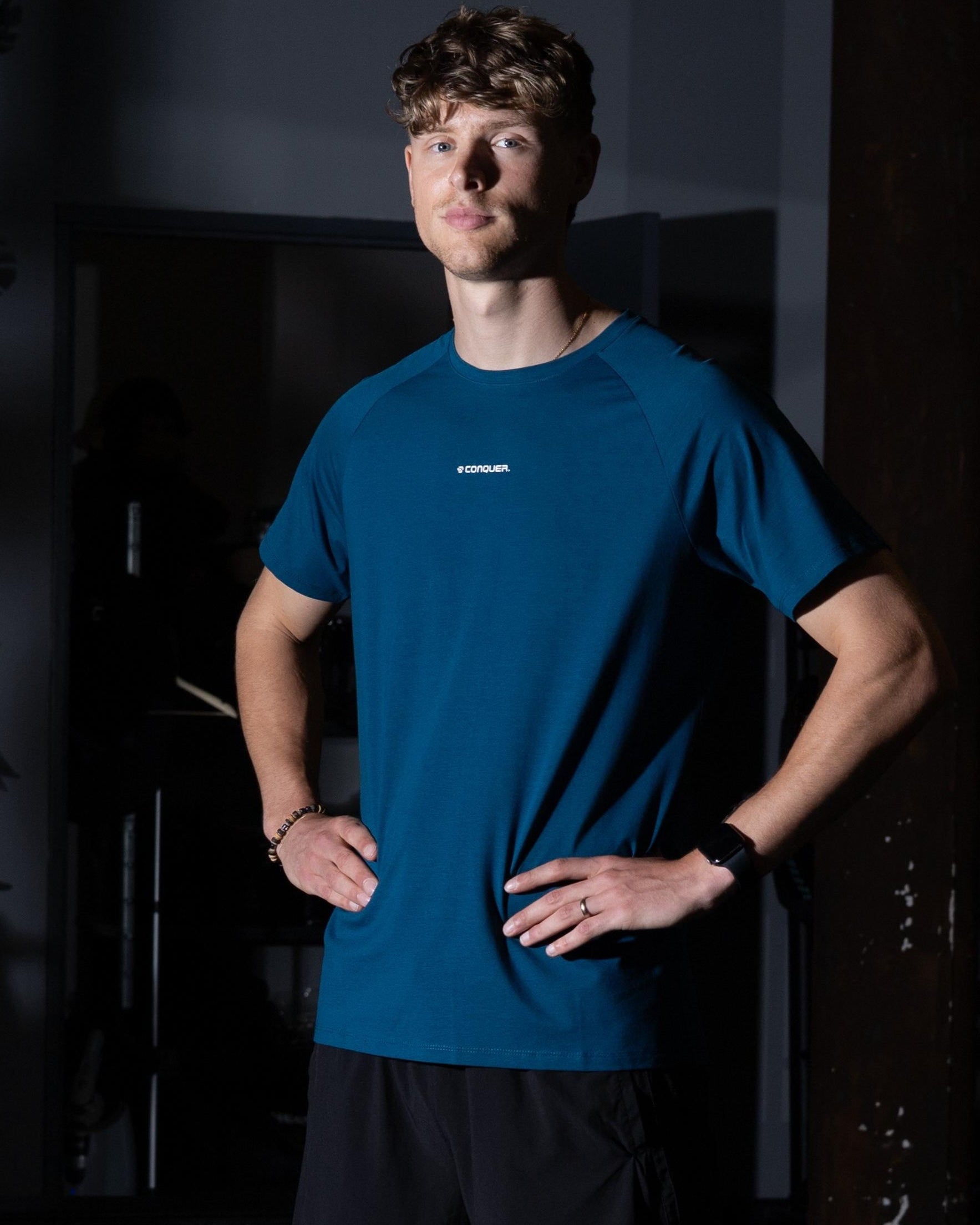 Man wearing a blue athletic shirt and black shorts standing in a dark room.