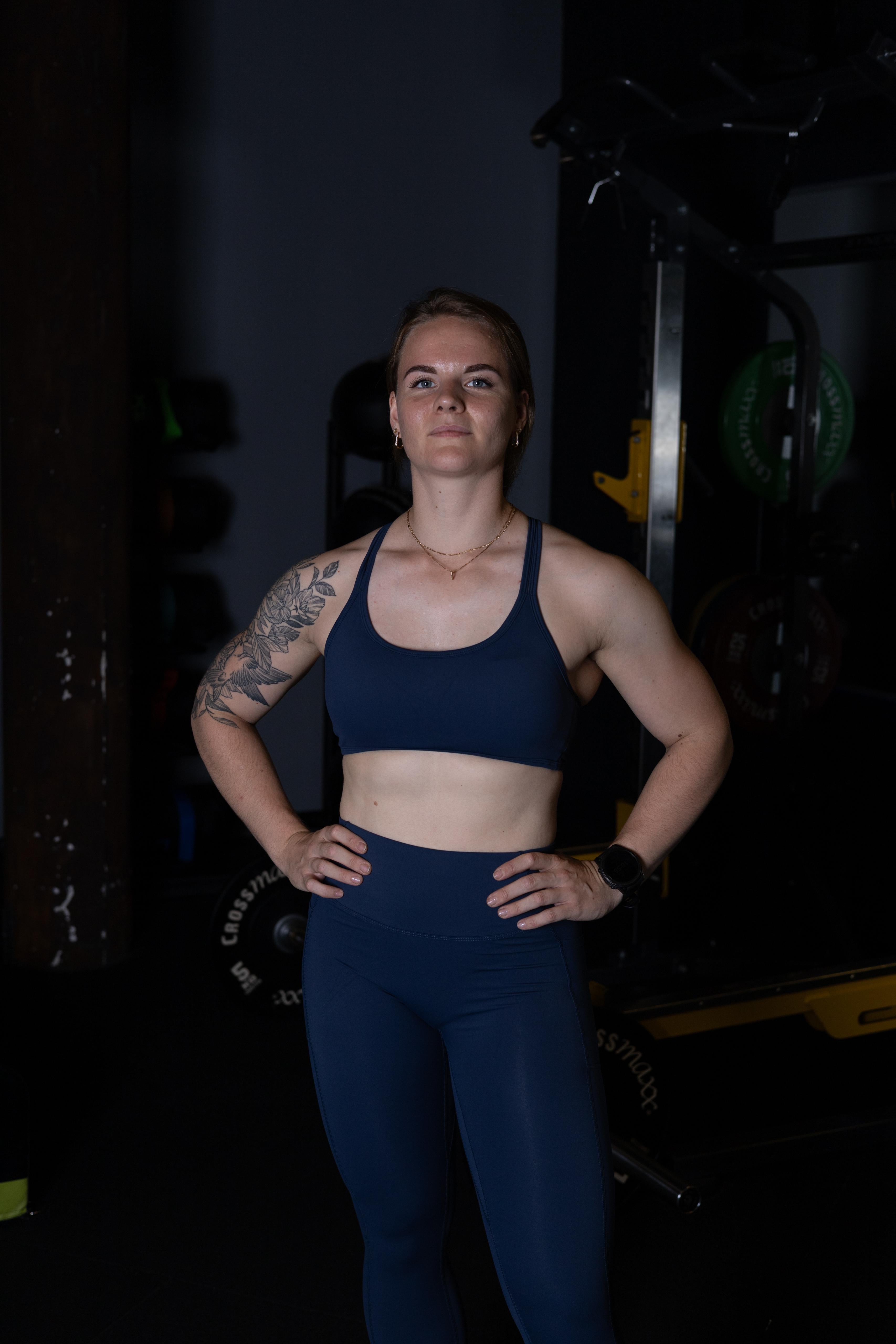 Woman in navy sports bra and leggings standing in a dark room with gym equipment.