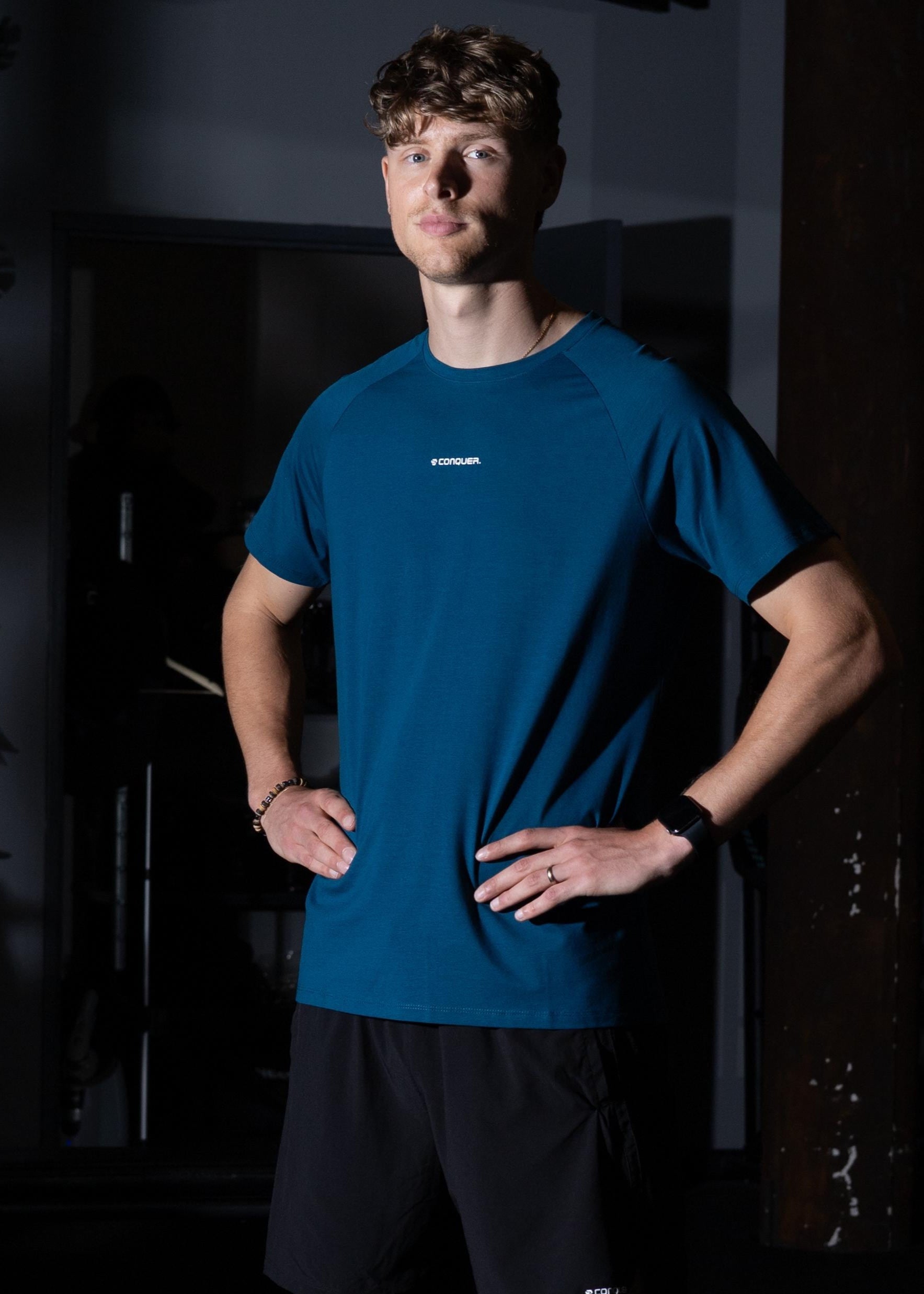 Man wearing a blue athletic shirt and black shorts standing in a dark room.
